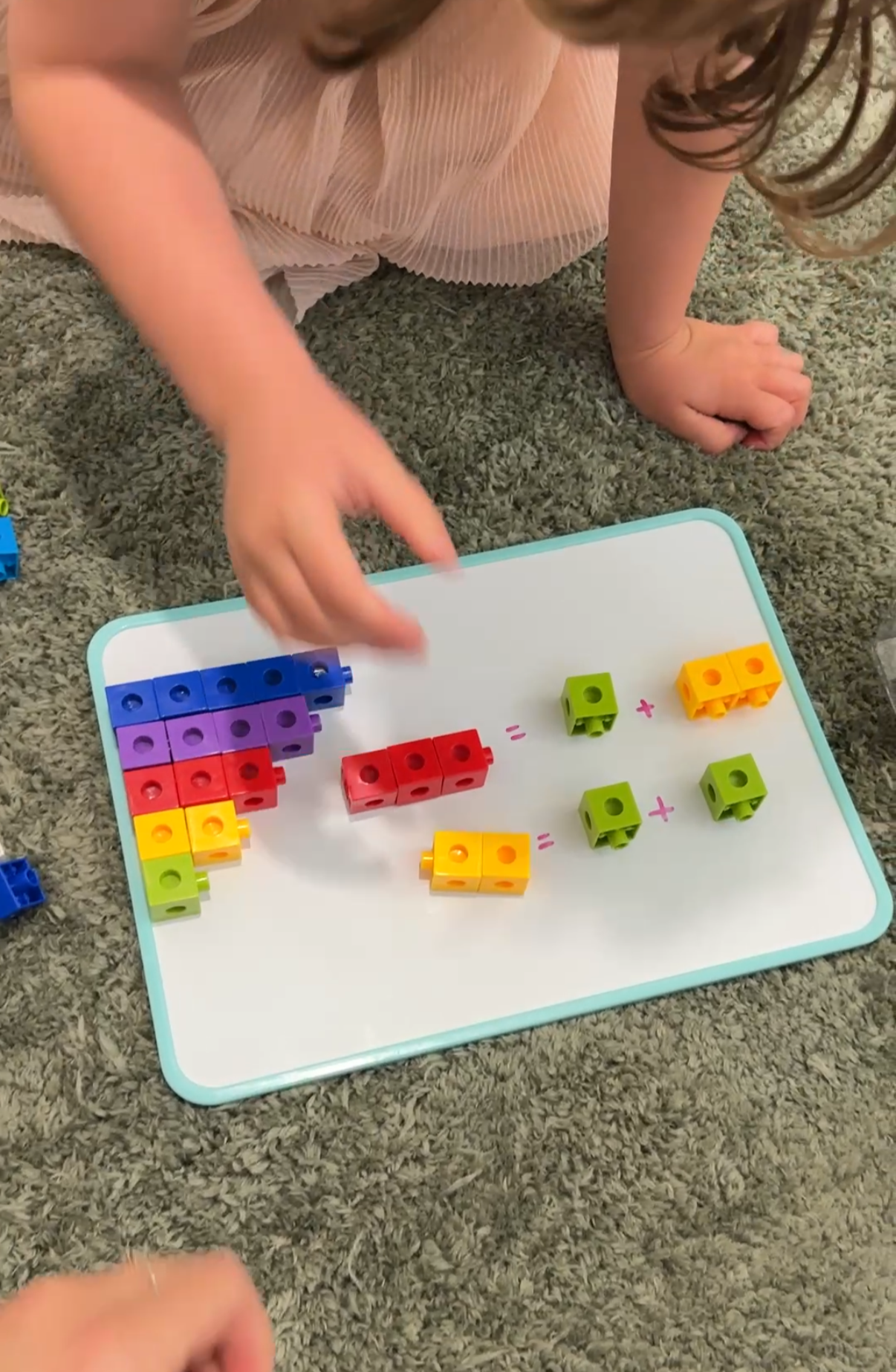 A child is playing with colorful building blocks on a whiteboard placed on a carpet. The blocks are arranged to illustrate basic addition, with some blocks grouped and mathematical symbols written on the board.