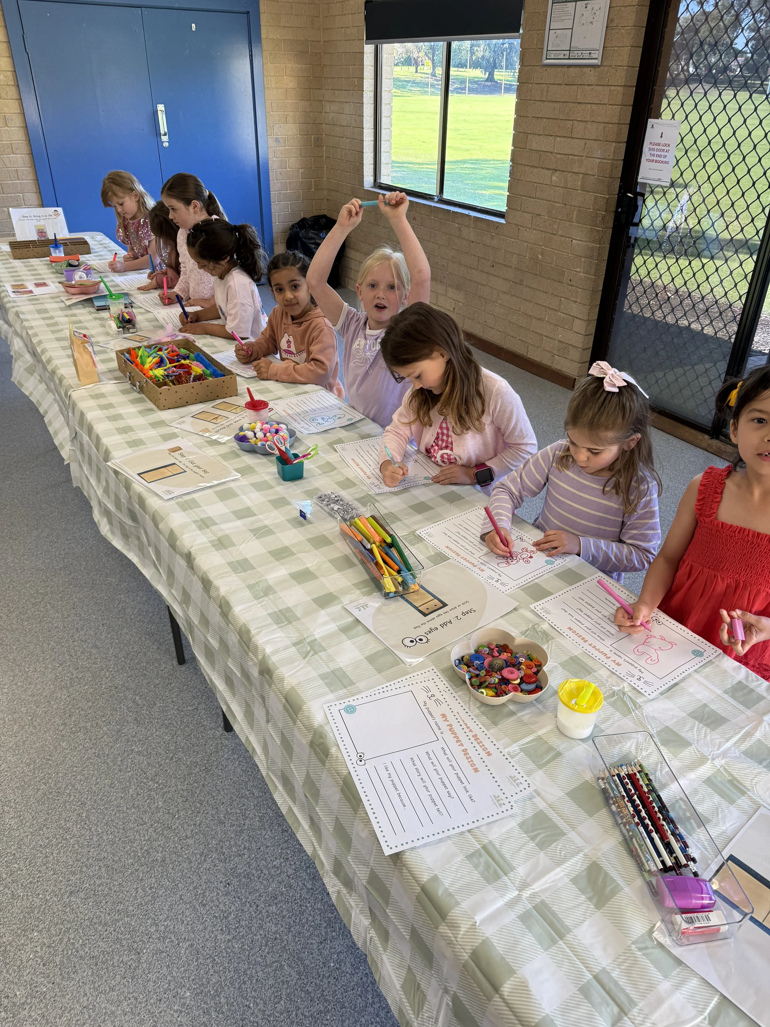 Young children sitting at a long table decorated with a checkered tablecloth, engaged in arts and crafts activities with markers, buttons, and paper, in a classroom with brick walls and large windows showing a grassy outdoor area.