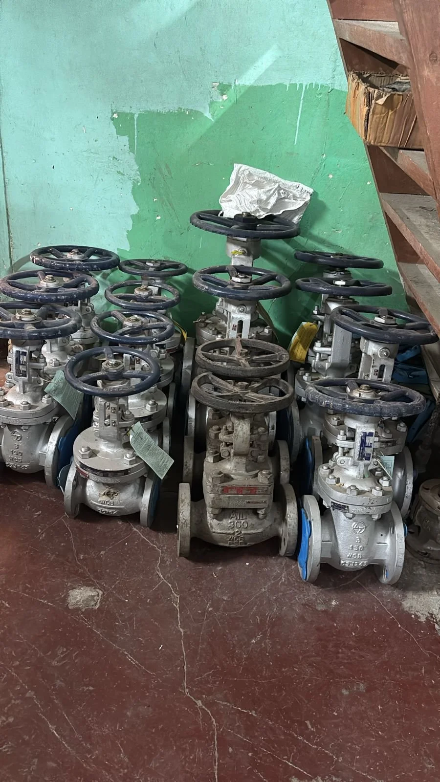 A group of industrial valves with wheel handles, arranged on the floor against a green wall and wooden shelving.