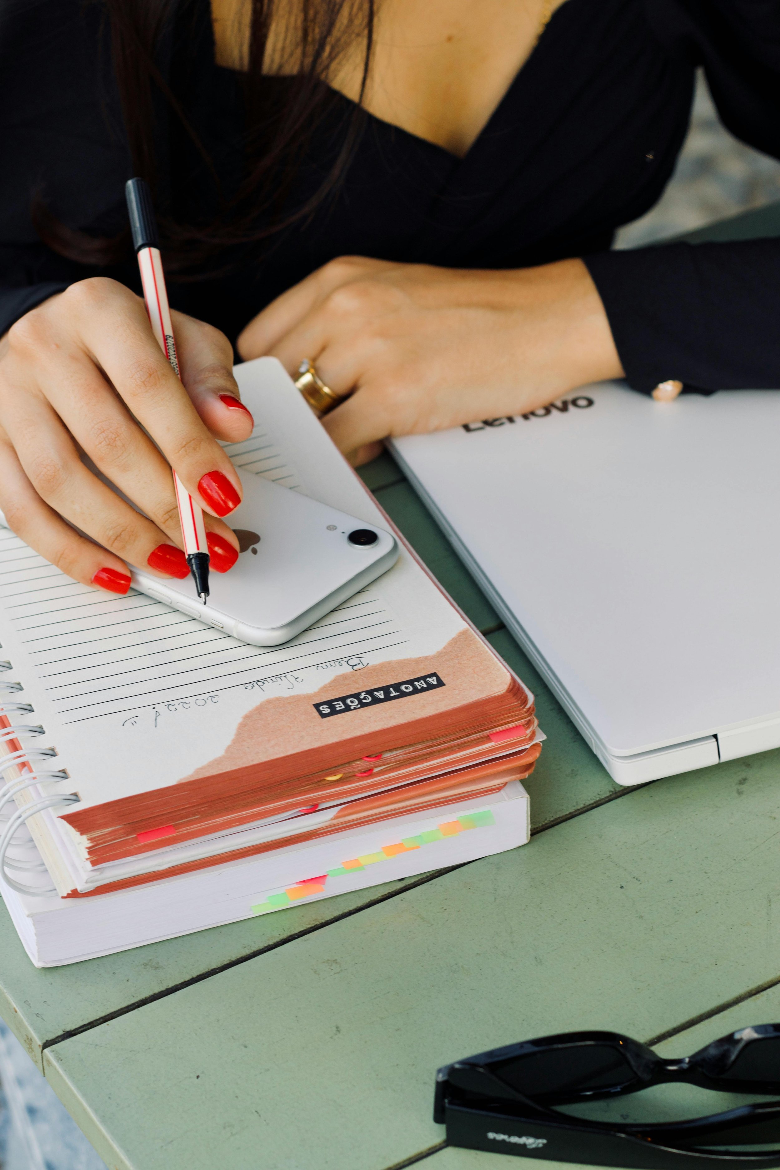 A person writing in a notebook with a pen, surrounded by a phone, a laptop, glasses, and a planner on a green desk.