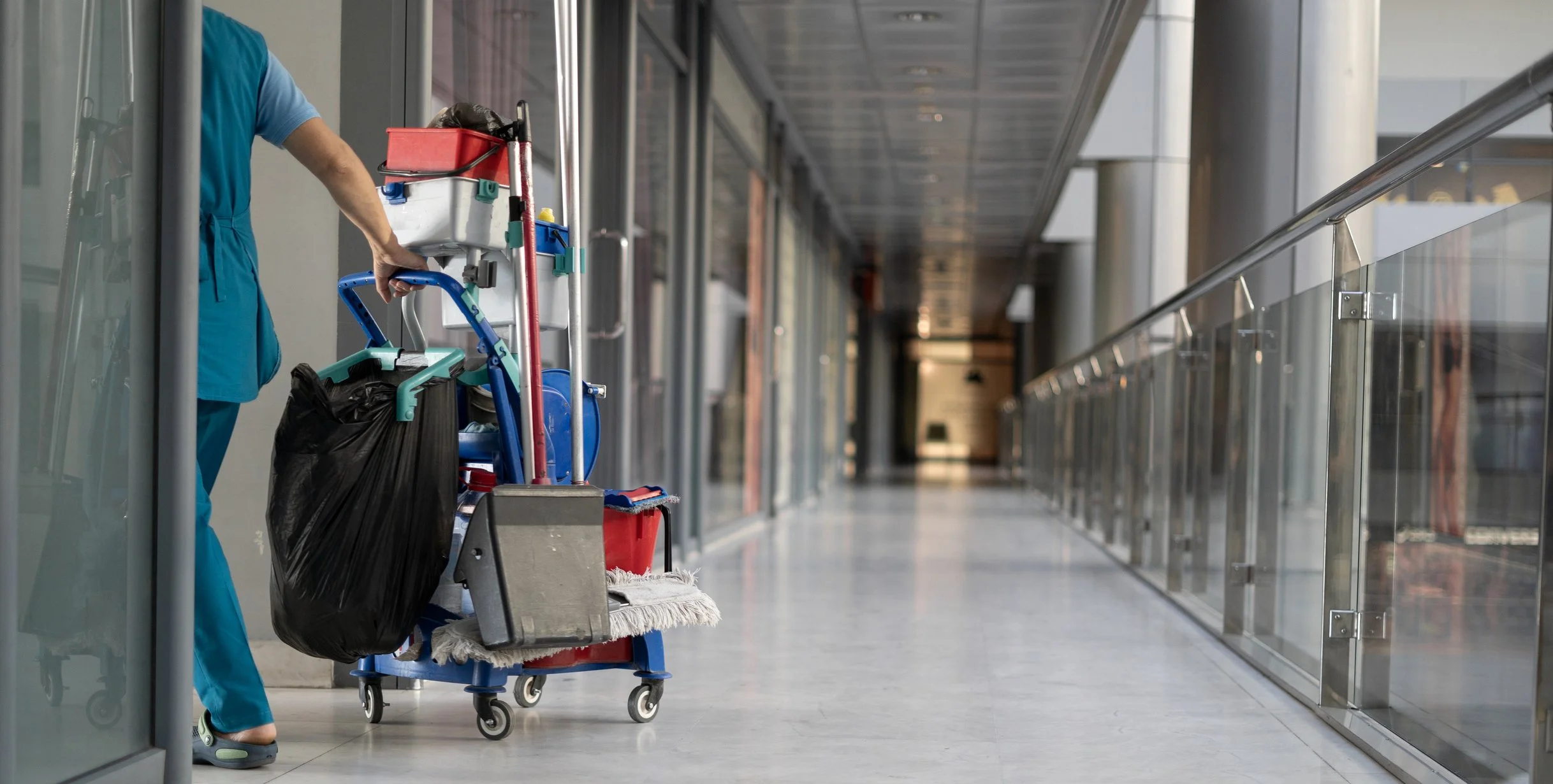 Janitor's cart with cleaning supplies and trash bag in an empty airport corridor.