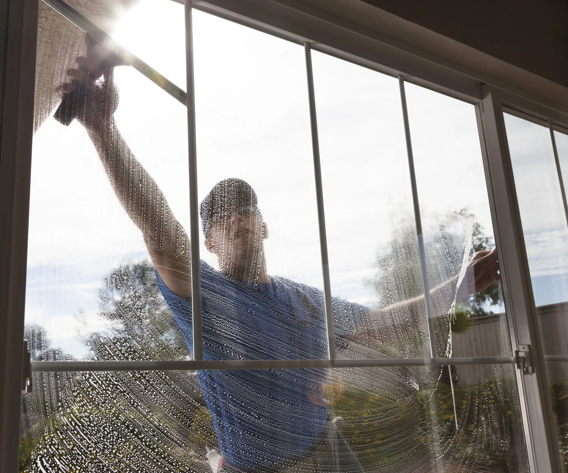 A person cleaning a large window with a squeegee, viewed from inside the house.