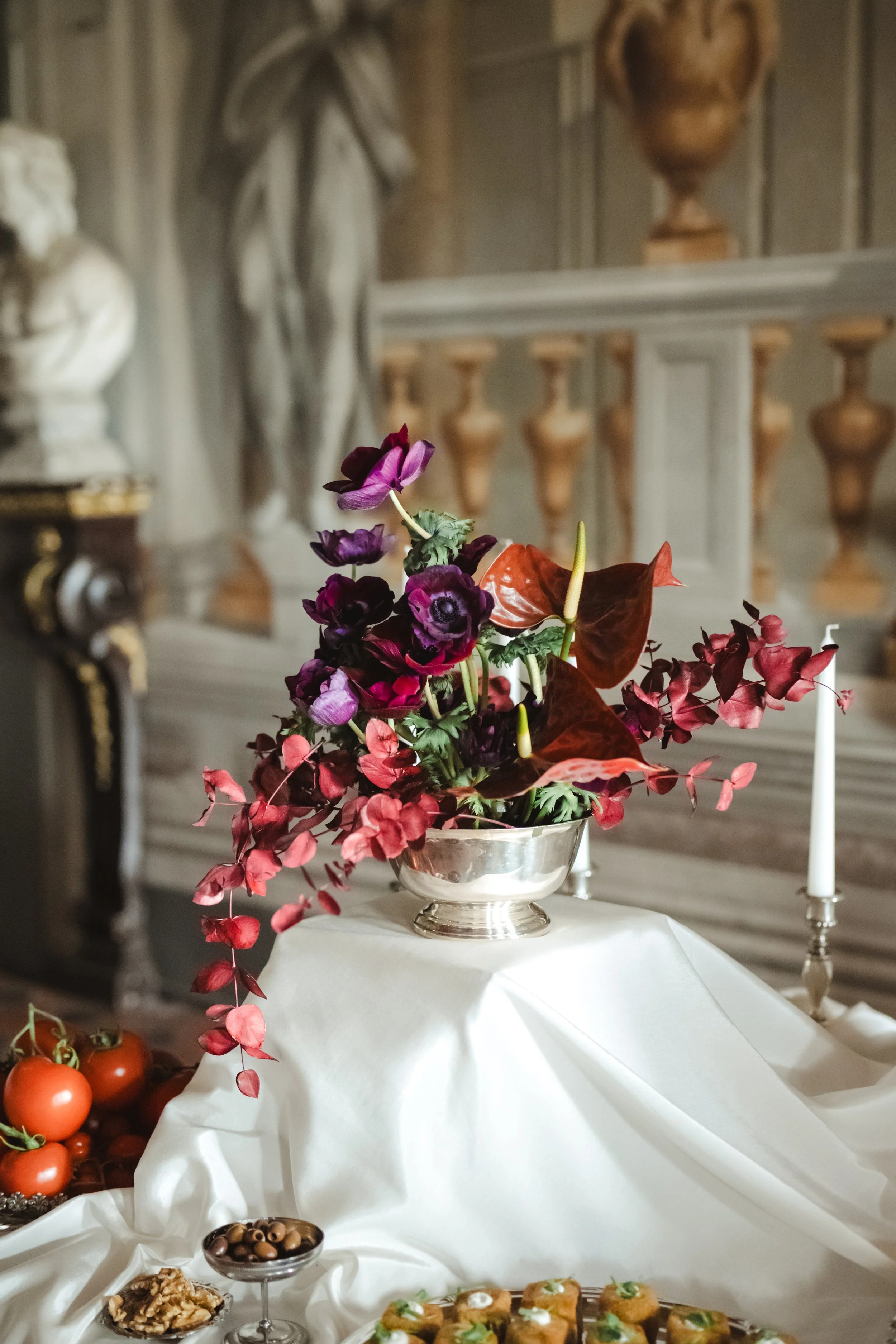 Vaso d'argento con un mazzo di fiori viola, rosso e verde su un tavolo ricoperto da una tovaglia bianca, con candelabro con candela bianca e altra decorazione e frutta sullo sfondo. Elisa Torrini - Etike Event Flower Design_ Firenze