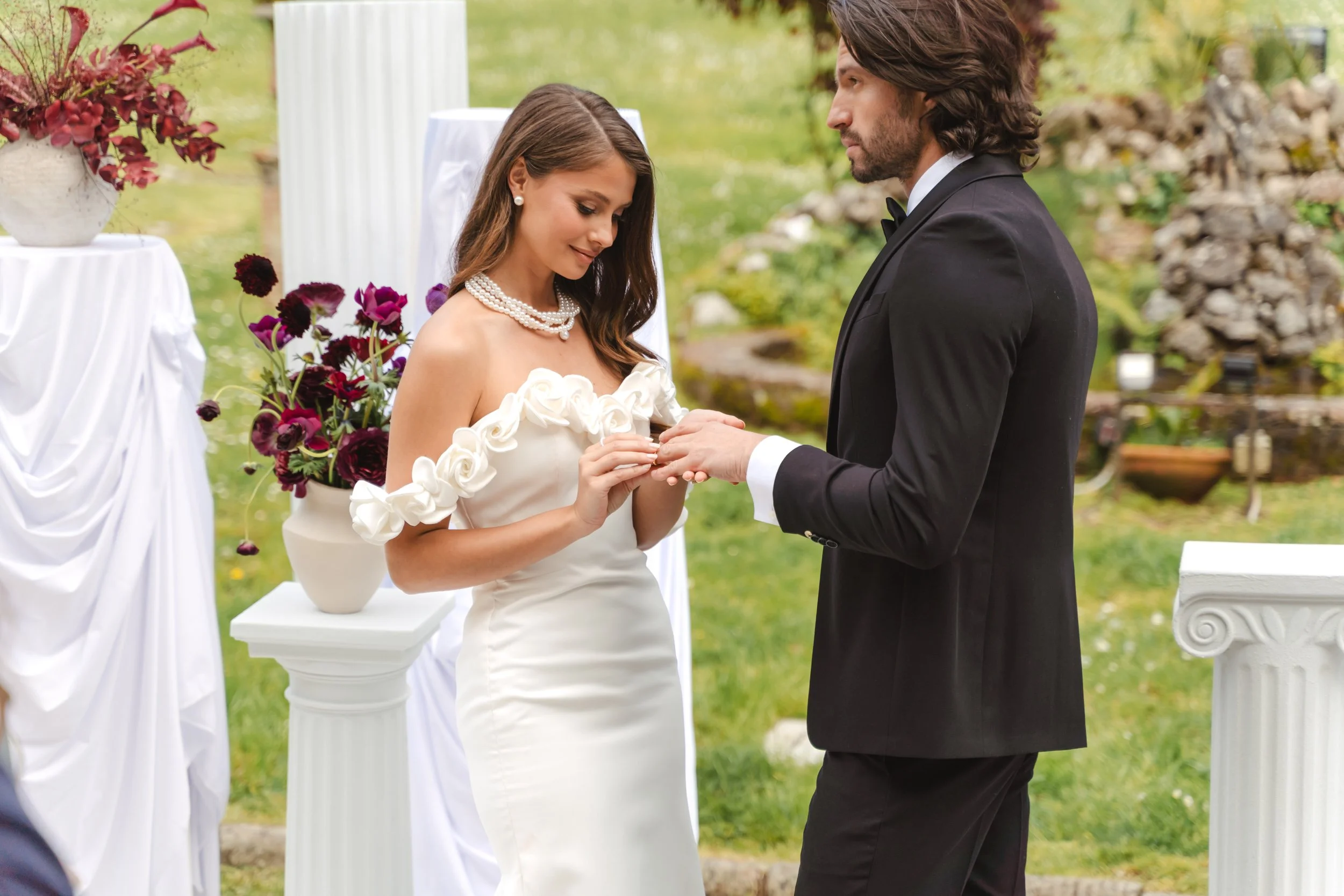 Coppia di sposi durante la cerimonia di matrimonio all'aperto, l'uomo sta mettendo l'anello alla donna, lei indossa un abito bianco con decorazioni di fiori, e una collana di perle, in un ambiente con decorazioni floreali e colonne. Elisa Torrini