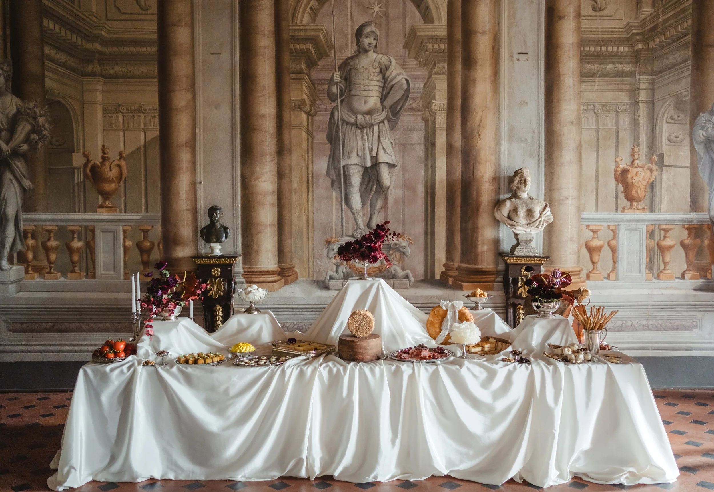 Una tavola imbandita con dolci e frutta, decorata con tovaglie bianche e fiori, davanti a un arazzo con statue romane e colonne. Elisa Torrini - Etike Event Flower Design_ Firenze