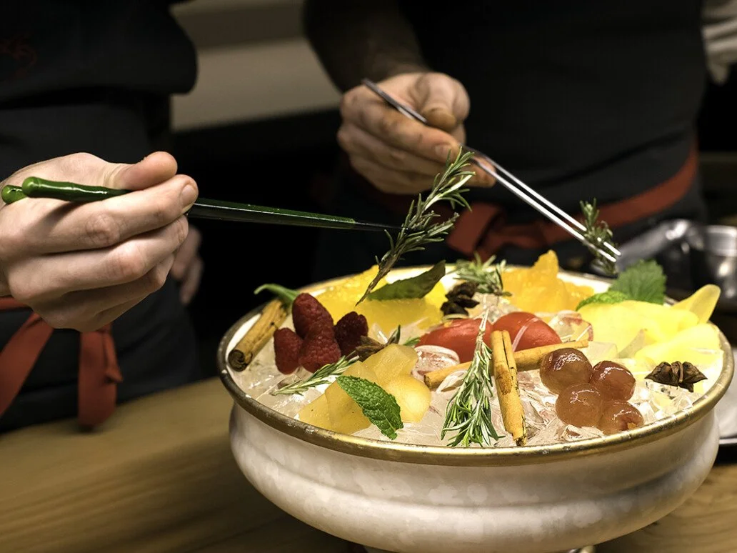 Persona decorando un plato con hierbas y frutas, como uvas, frambuesas y cítricos, en un ambiente de cocina.