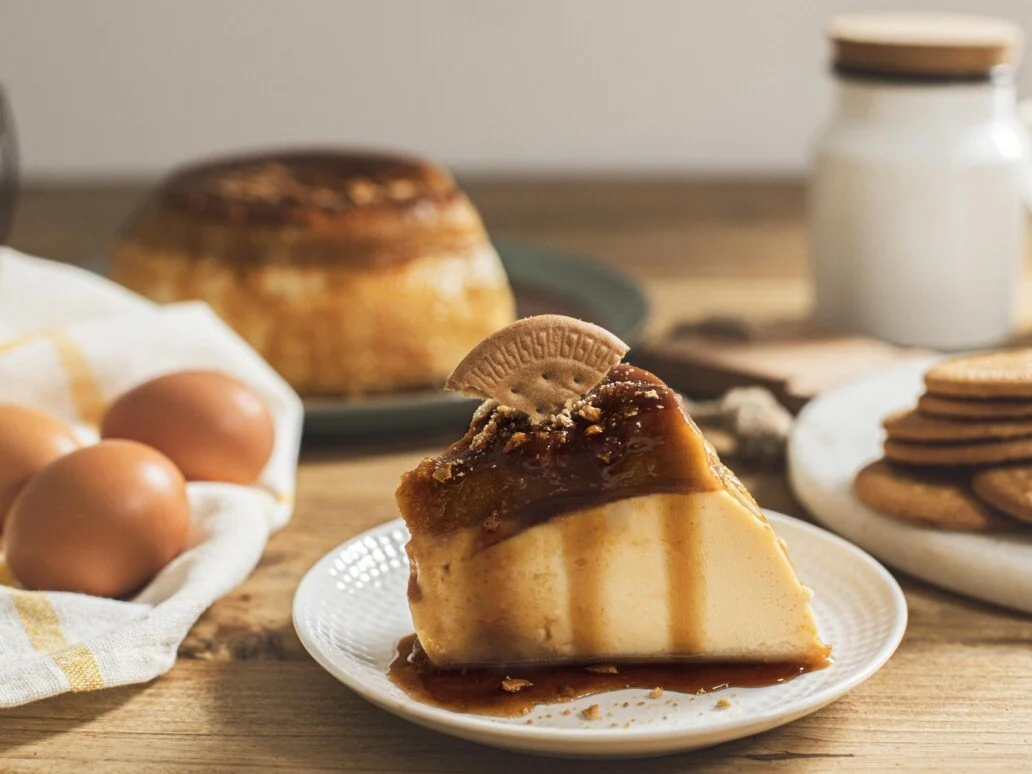 Porción de pay de vainilla con caramelo y galleta encima, en un plato blanco sobre una mesa de madera, con huevos, una torta y galletas a su alrededor.