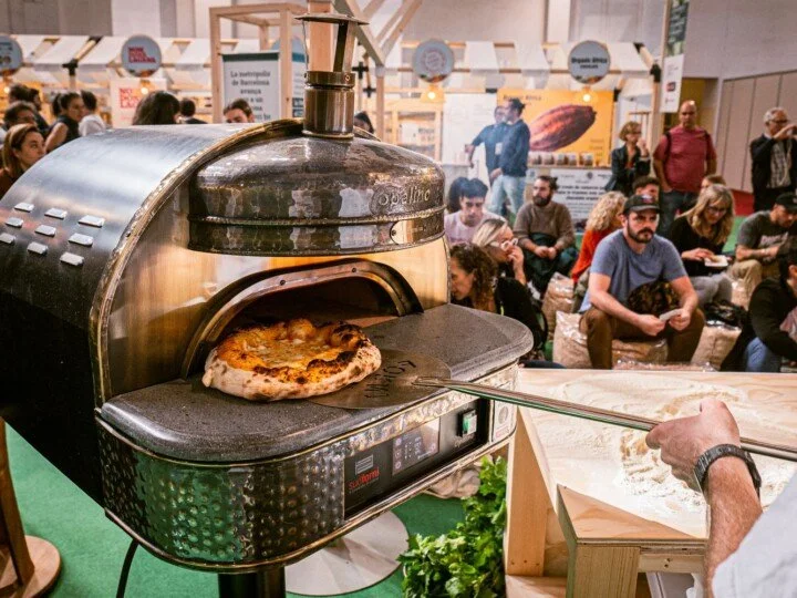 Persona sacando una pizza del horno de leña en una feria o evento, con muchas personas sentadas y de pie en el fondo.