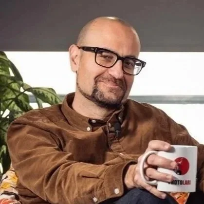Hombre calvo con barba y gafas, sosteniendo una taza, sentado en una habitación con plantas y ventana de fondo.