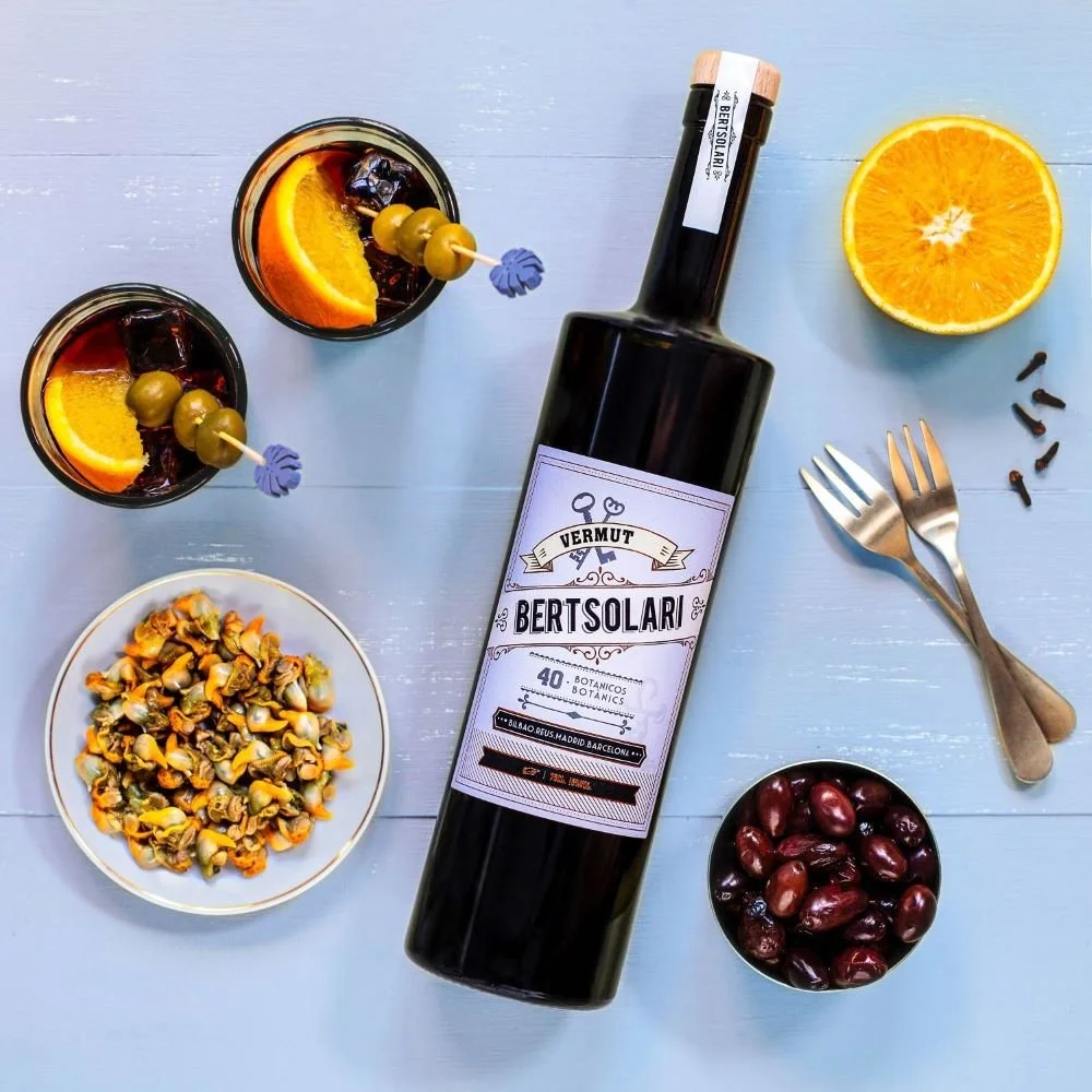 Botella de vermut Bertsolar, dos copas con bebida, rodajas de naranja, aceitunas, una fruta partida, una taza con mezcla de frutos secos, unos granos de pimienta y tenedores sobre una mesa de color azul.