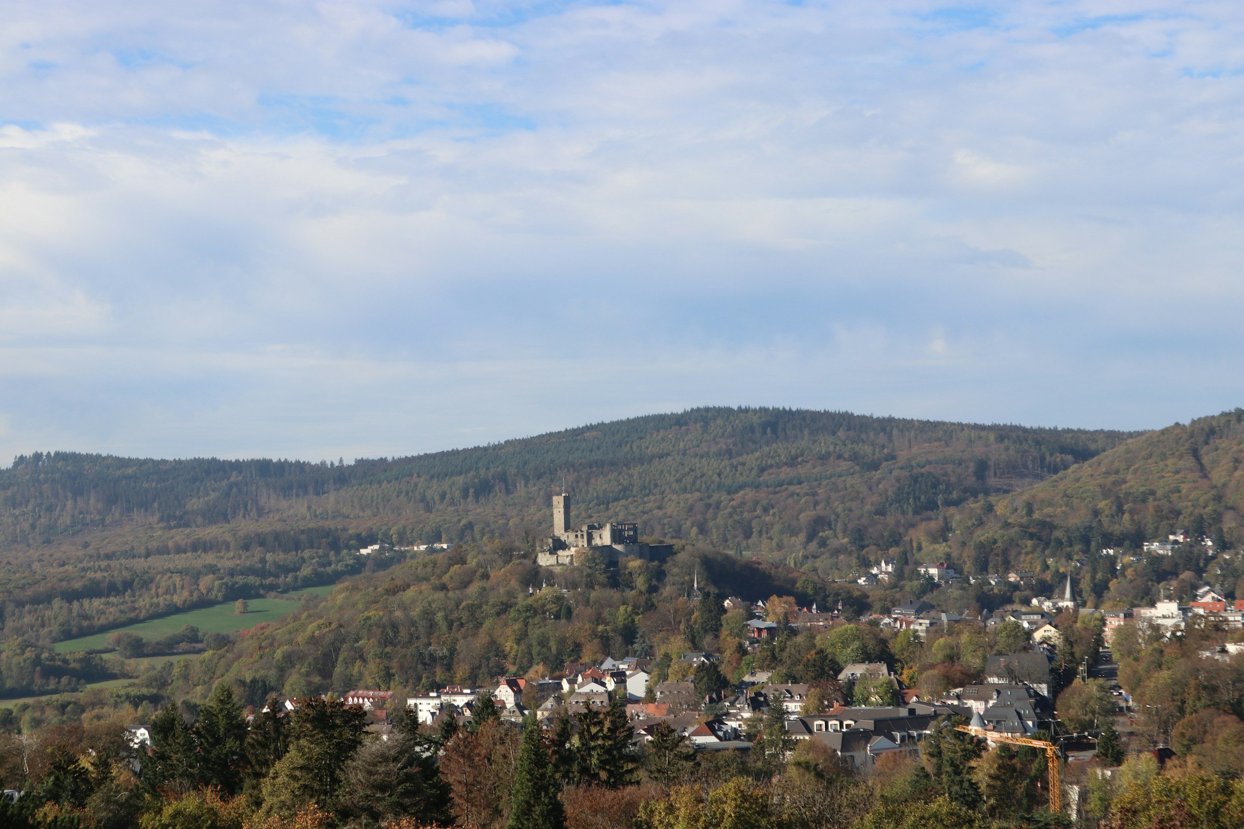 Blick auf eine kleine Stadt mit einem Schloss auf einem Hügel, umgeben von Wäldern und Hügeln, bei sonnigem Himmel mit wenigen Wolken.