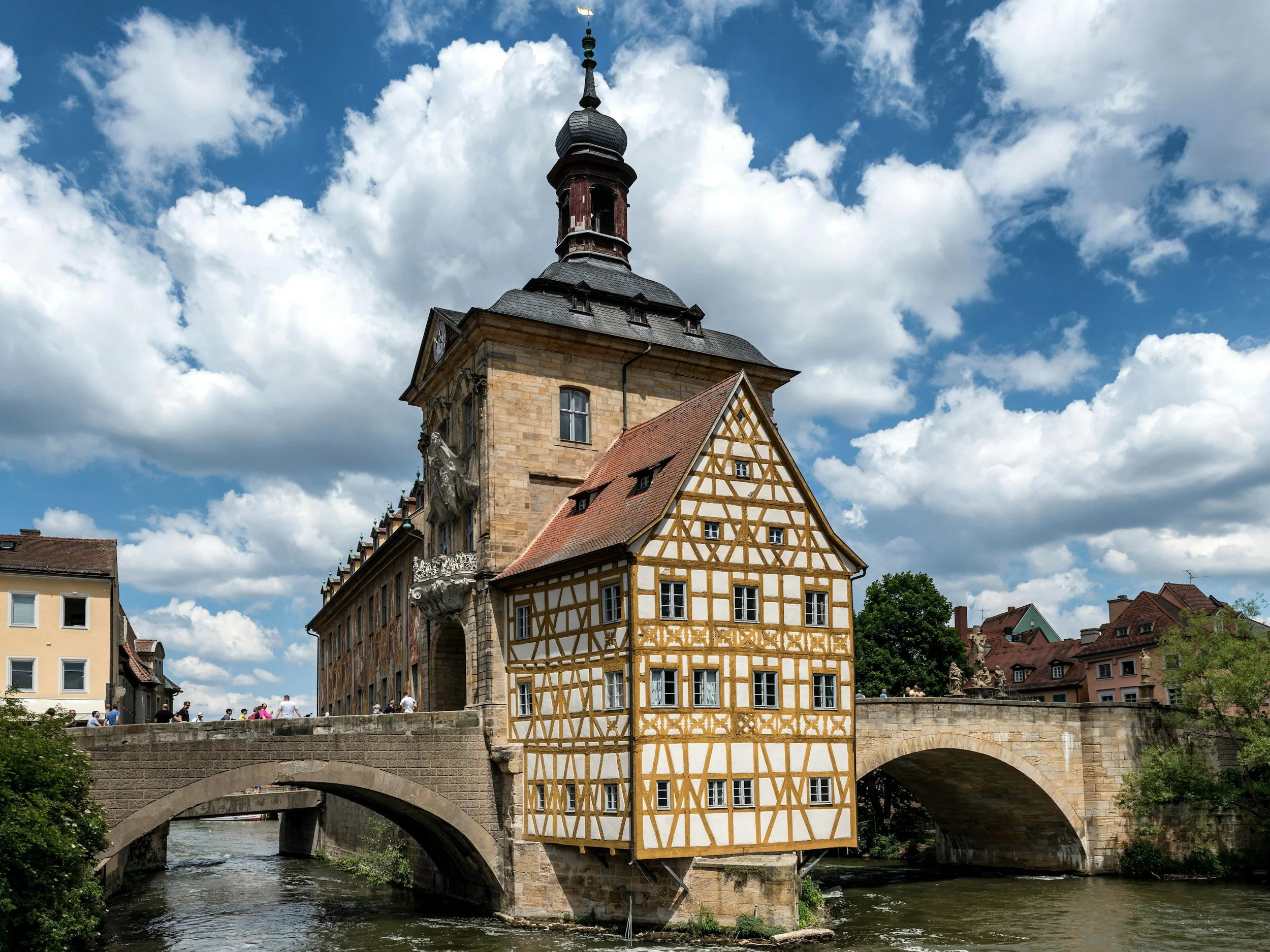 Bamberg: Historisches Gebäude mit Fachwerkfassade und Turm über einem Brückenbogen, am Fluss in einer deutschen Stadt, bei sonnigem Himmel mit Wolken.