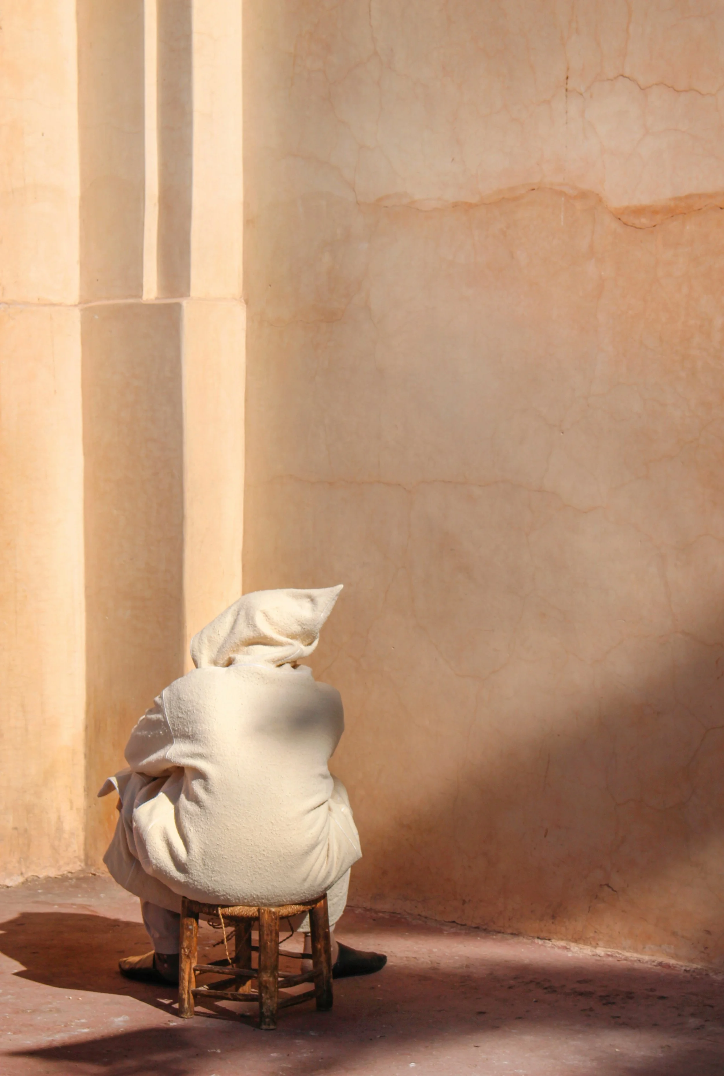 Personne assise sur un petit tabouret en bois, portant des vêtements amples de couleur clair, avec un foulard ou un chapeau sur la tête, dans un environnement avec des murs de couleur beige ou ocre et un sol en terre. La personne est vue de dos.