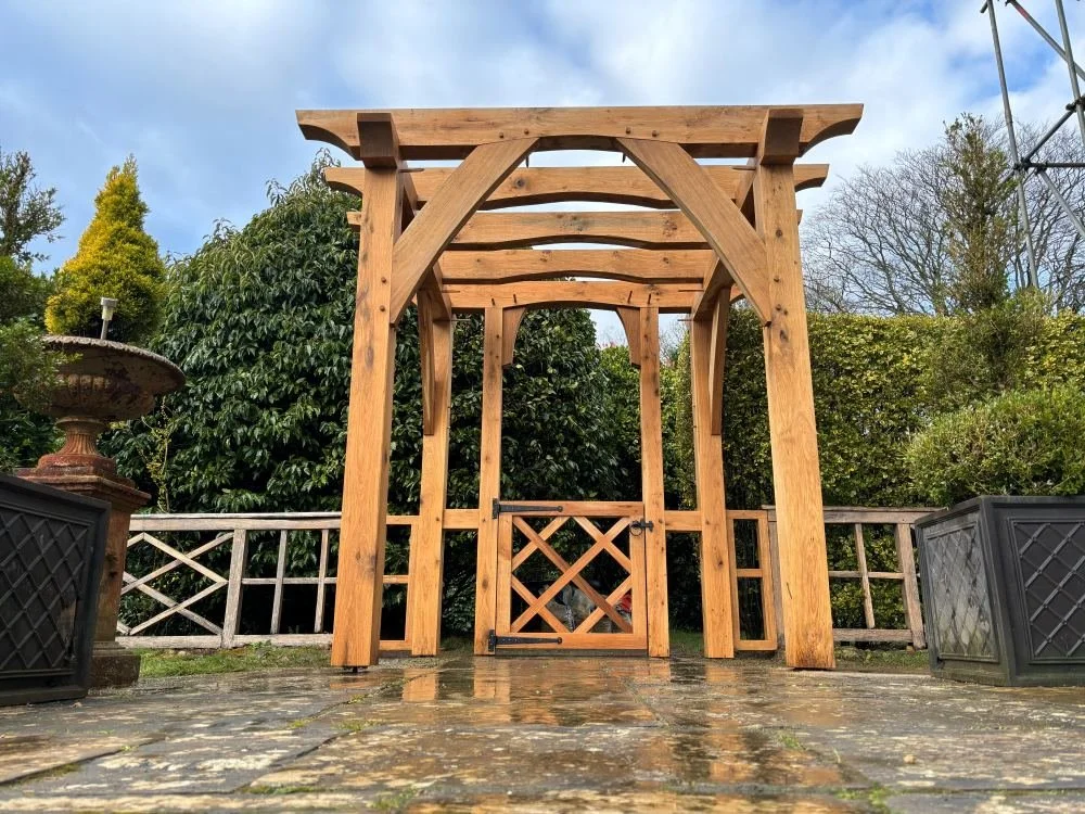 A newly constructed wooden pergola in a garden area with a stone pathway, surrounded by bushes and trees, with a partly cloudy sky overhead.