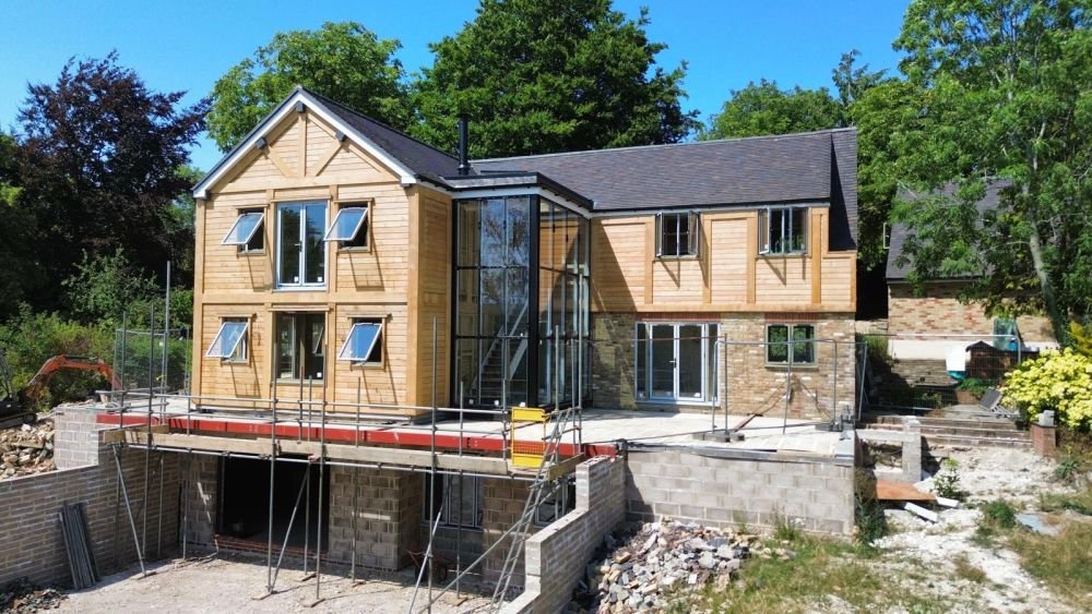 Under construction house with wooden and brick exterior, scaffolding, and a glass-walled staircase connecting levels, surrounded by trees and construction materials.