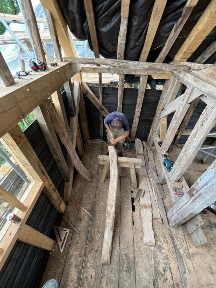 A person working on a wooden construction inside a partially built structure, with exposed wooden beams and framing, tools, and building materials around.
