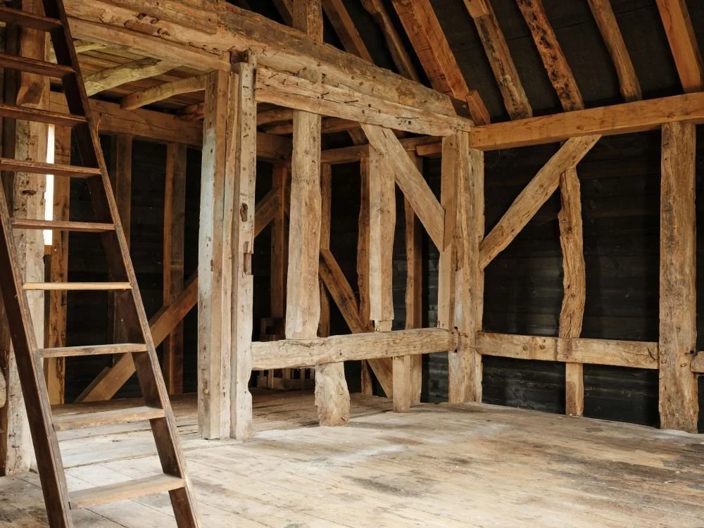 The interior of an unfinished wooden attic with exposed beams and a wooden ladder leaning against the wall.