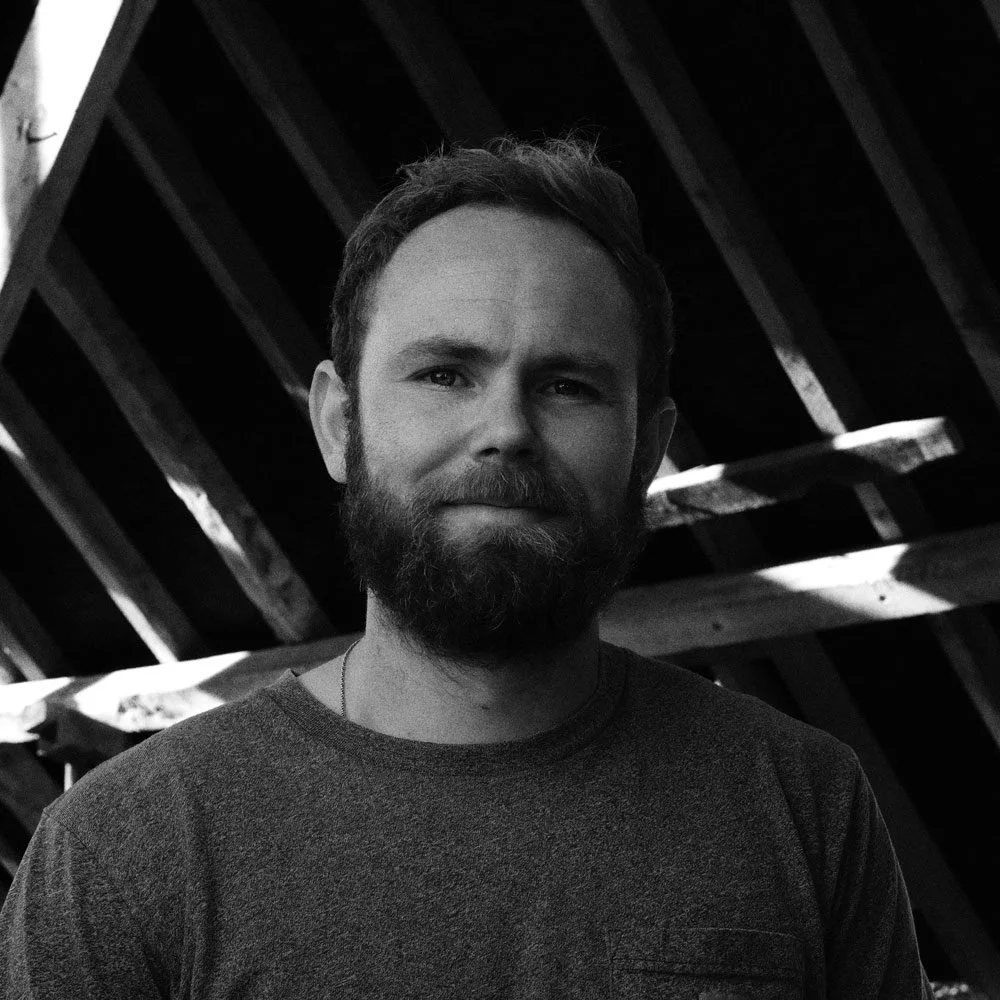 A man with a beard and short hair posing in a wooden structure with slanted beams, in black and white.