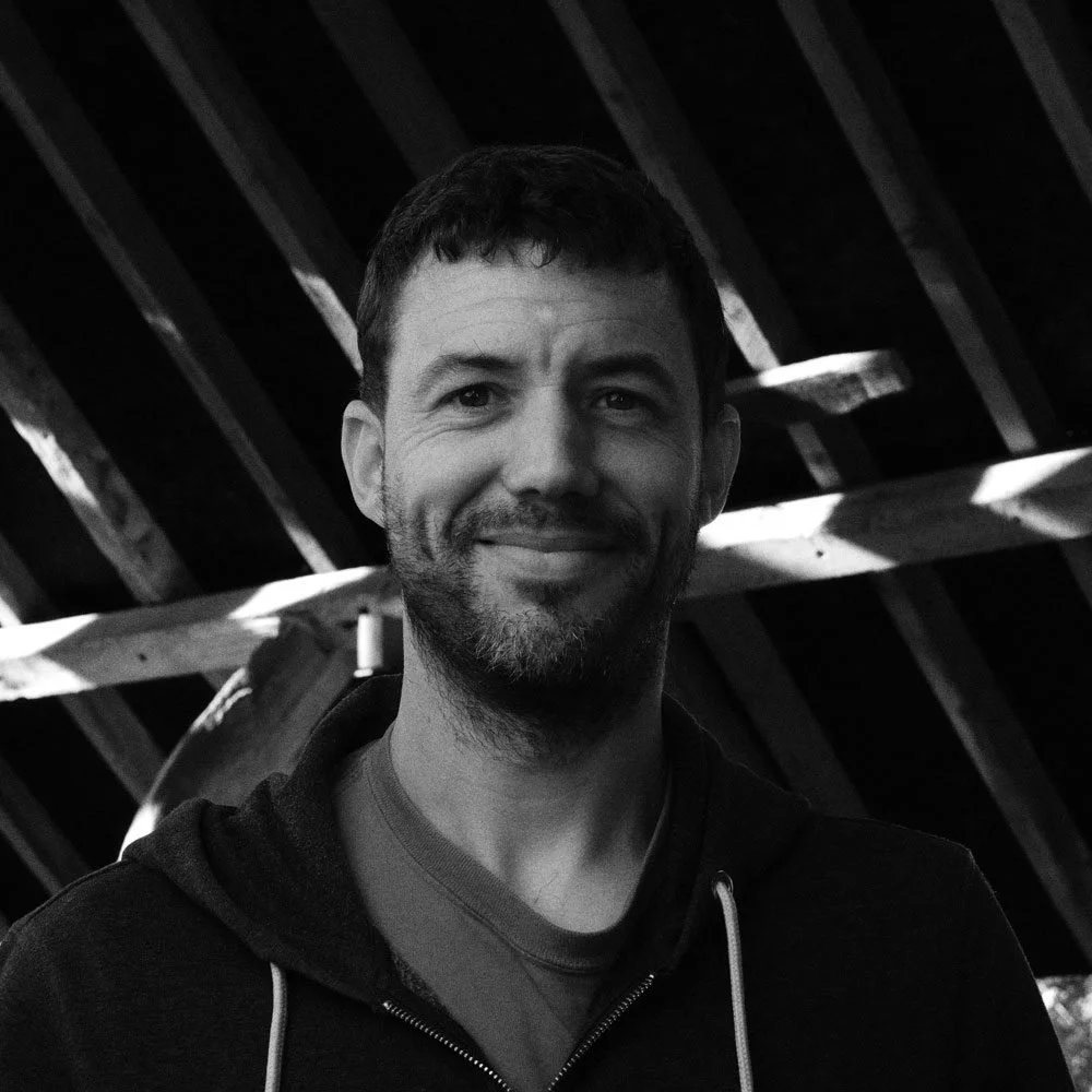 Black and white photo of a smiling man with short dark hair and a beard, standing outdoors under a wooden structure.