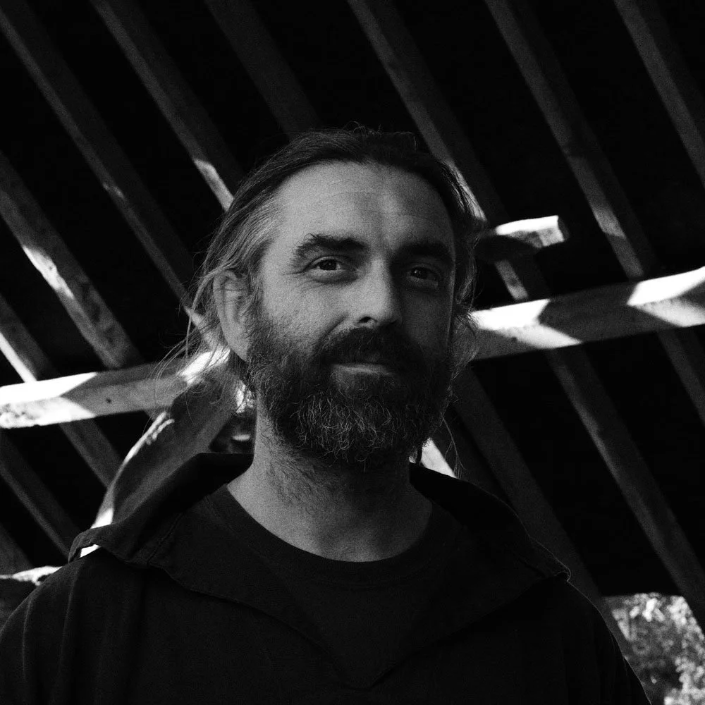 A black and white photo of a man with long hair and a beard, smiling slightly, with a wooden roof structure in the background.