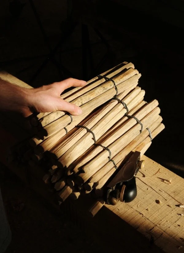 A person's hand holding a bundle of labeled wooden sticks tied together with black string on a workbench, with a hand plane nearby in a woodworking shop.