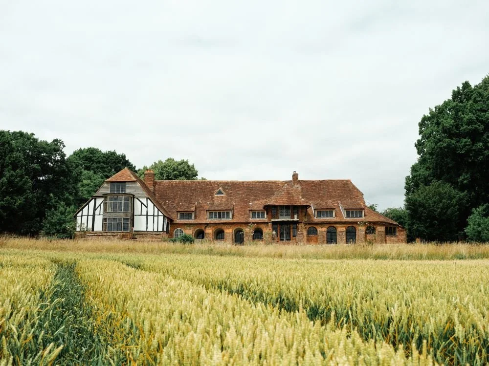 An old brick house with a red-tiled roof, surrounded by a field of wheat or tall grass, and trees in the background.