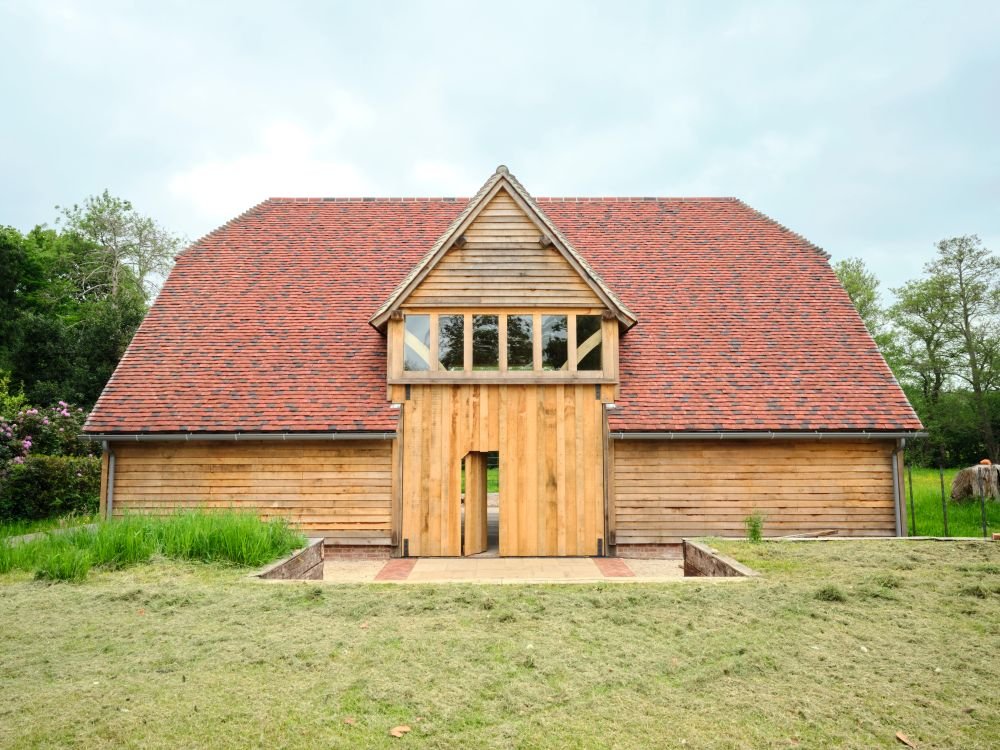 A wooden barn with a steep red shingle roof and a central entrance with an upper windowed dormer.