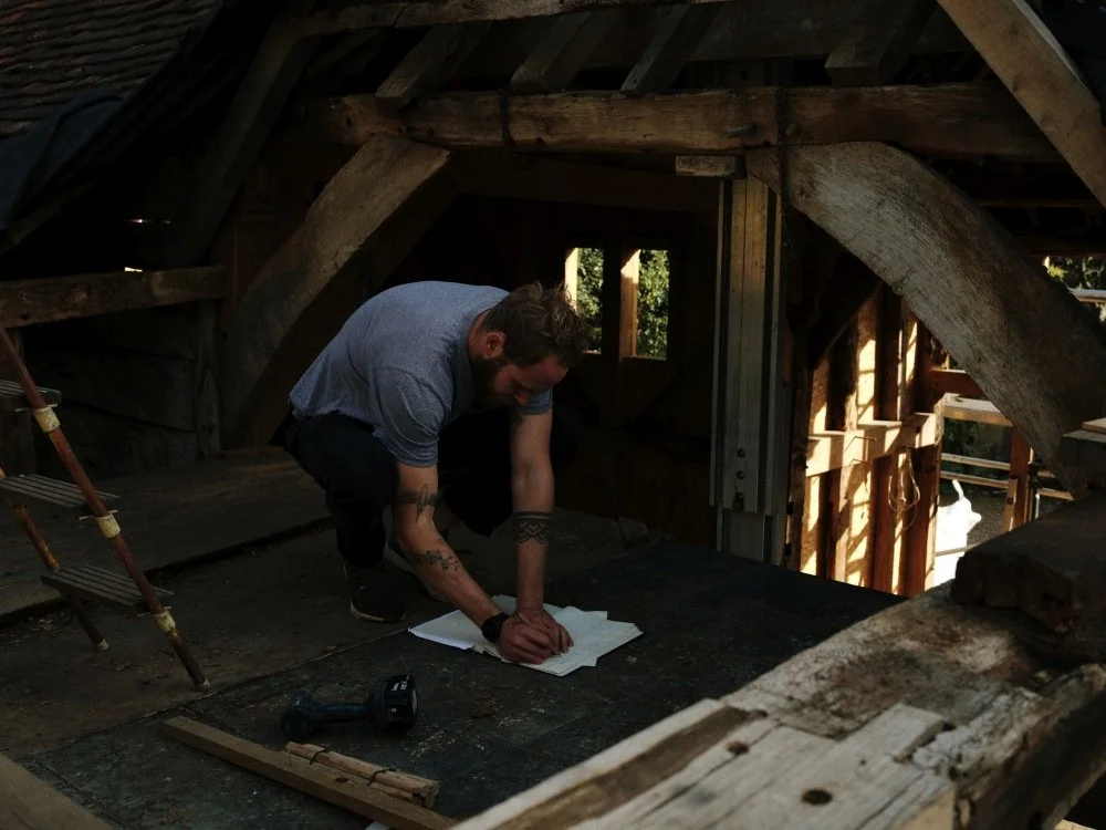 A man inside a rustic attic or loft space, kneeling on the floor and writing or drawing on a piece of paper. The space has wooden beams, some of which are curved and aged, and a small window in the background. Tools and a ladder are also visible.