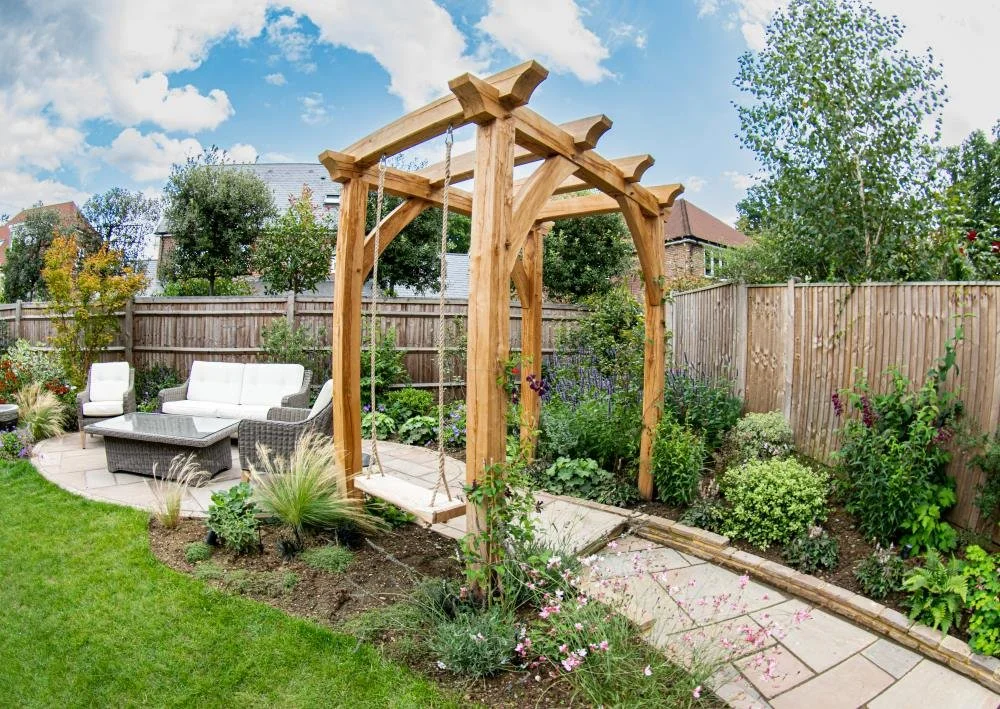 A backyard garden with a wooden swing, garden furniture, and various plants and shrubs, enclosed by a wooden fence under a partly cloudy sky.