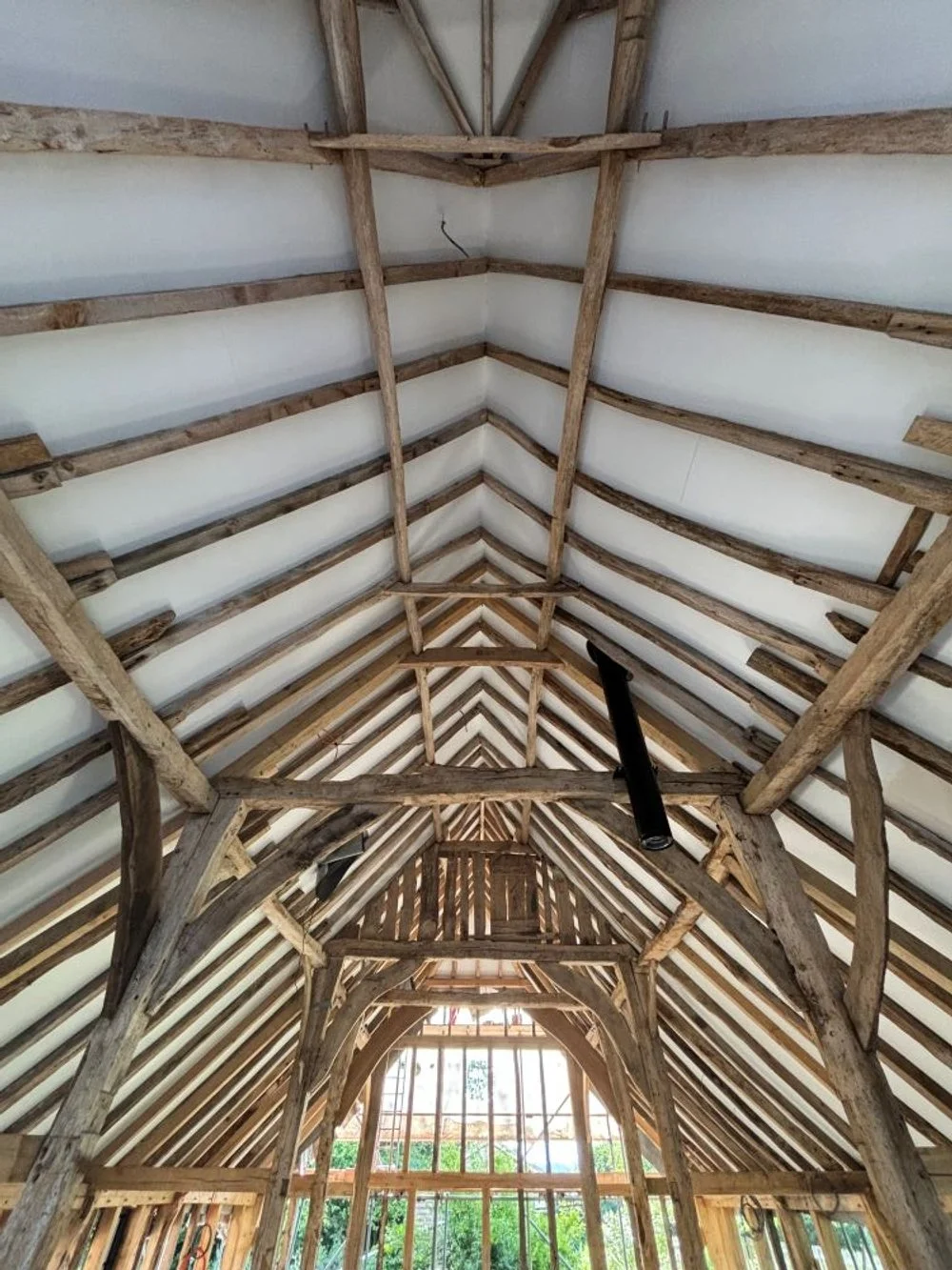 View of the interior of a barn under construction, showing wooden beams supporting the roof structure. The roof is lined with insulation material, and sunlight is visible through the open windows at the far end.