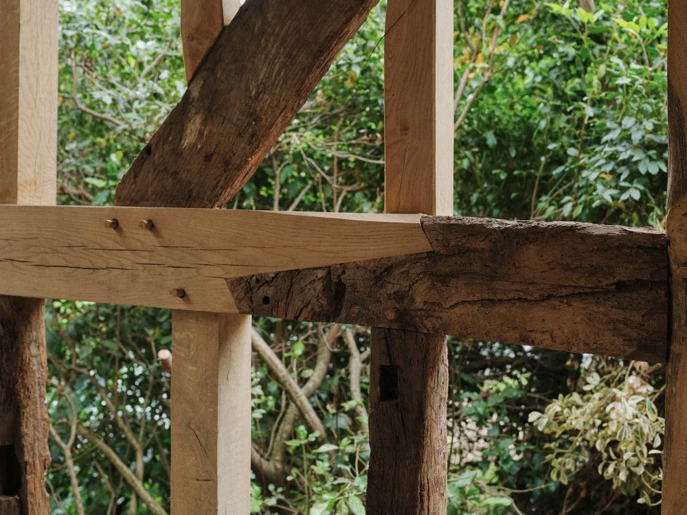 Close-up view of a wooden structure made of rough and smooth wooden beams, with a background of green foliage and tree branches.