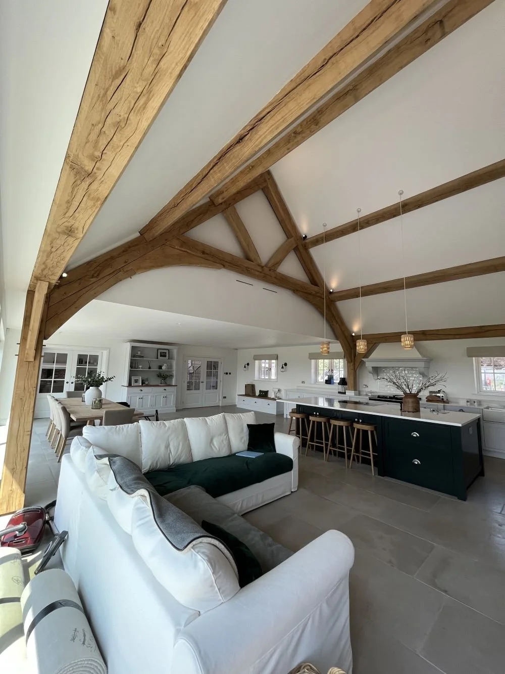 Open-concept living room with vaulted ceiling featuring exposed wooden beams, white walls, a white sofa with black and gray pillows, a large kitchen island with green cabinets, wooden bar stools, and dining area in the background.