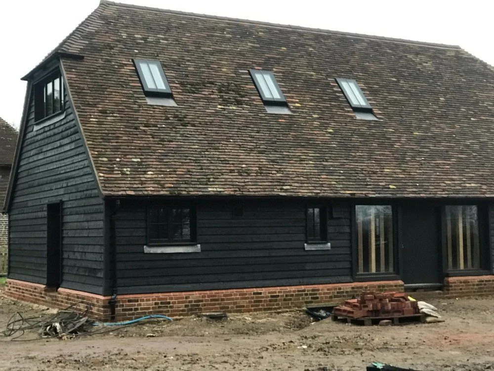 A house under construction with black wooden siding, new windows, and a brick foundation. There are construction materials and tools on the ground.