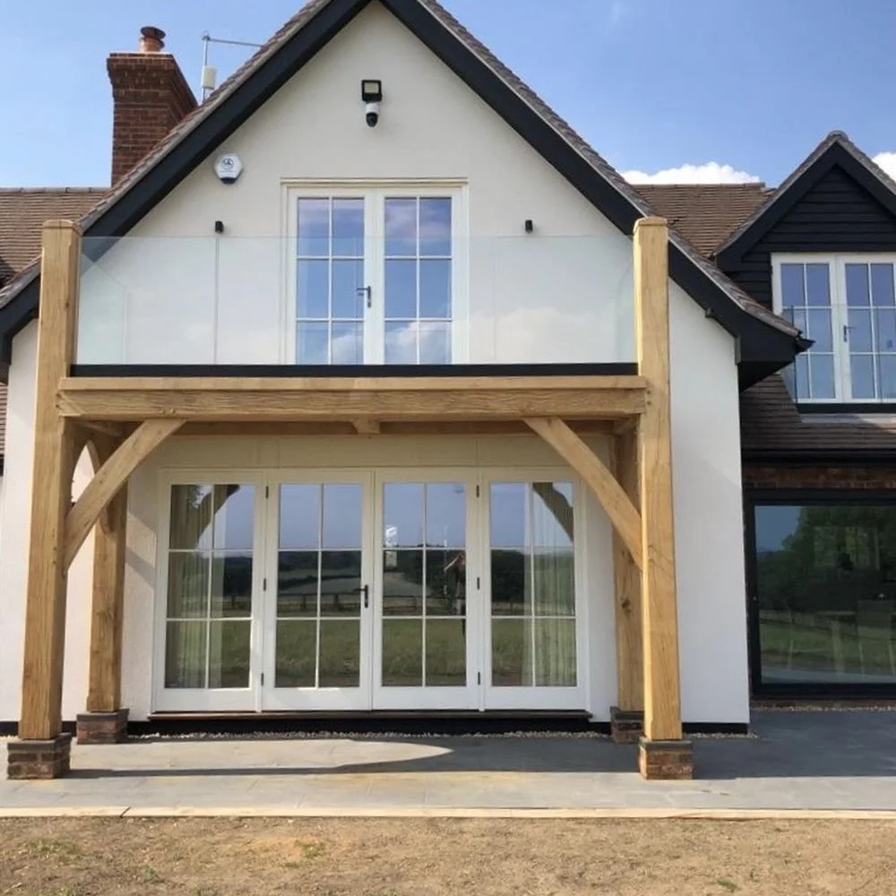 A two-story house with a glass balcony on the upper floor and large glass doors on the ground floor. The house has a sloped roof, and there are wooden supports framing the ground-level doors. The upper balcony has glass panels with wooden posts, and there is a smoke vent on the roof.