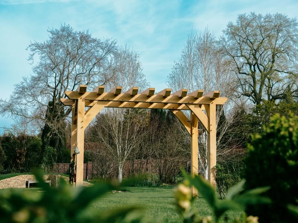 A wooden garden arbor with a lattice top in a landscaped yard, surrounded by trees and shrubs.