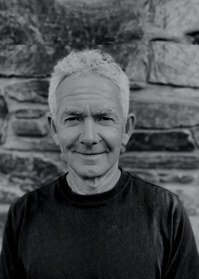 Black and white photo of an older man with gray hair, wearing a dark t-shirt, standing outdoors in front of a rock wall.