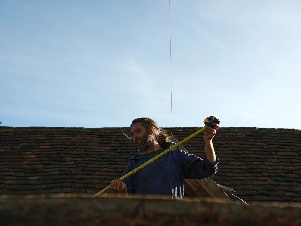 A man with long hair and a beard measuring the roof of a house with a yellow tape measure during daylight.