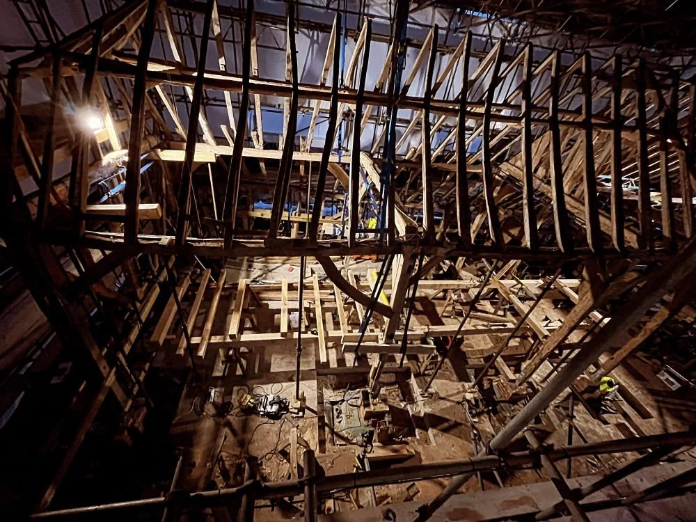 Interior view of a construction site showing wooden framework and scaffolding for a large structure, with workers in safety gear.