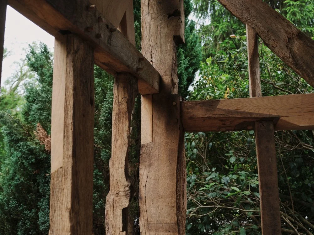Close-up of a wooden structure under construction, showing vertical and horizontal rough-hewn wooden beams and planks with joins and nails, with green foliage in the background.