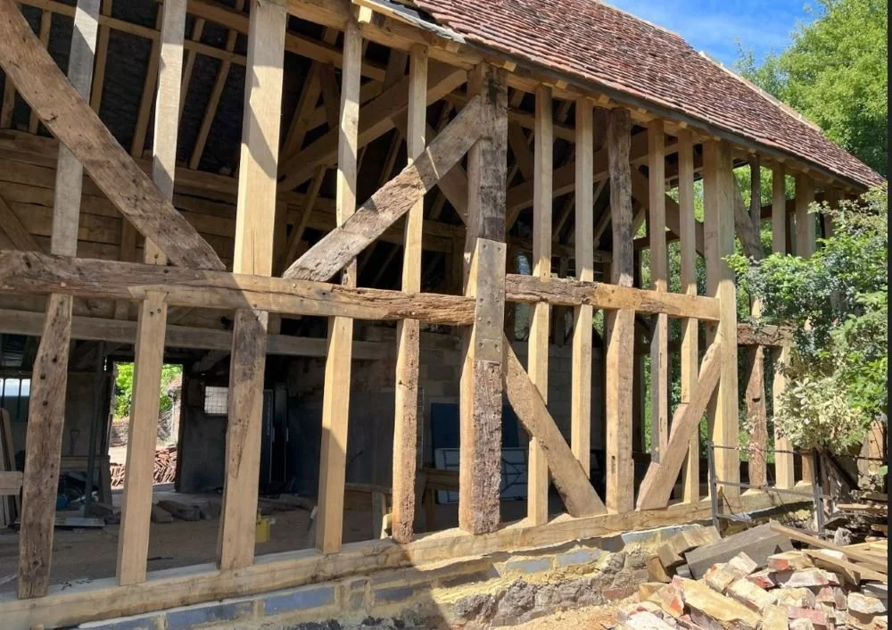 Wooden framing for a house under construction, with visible roof and support beams, and construction materials on the ground.