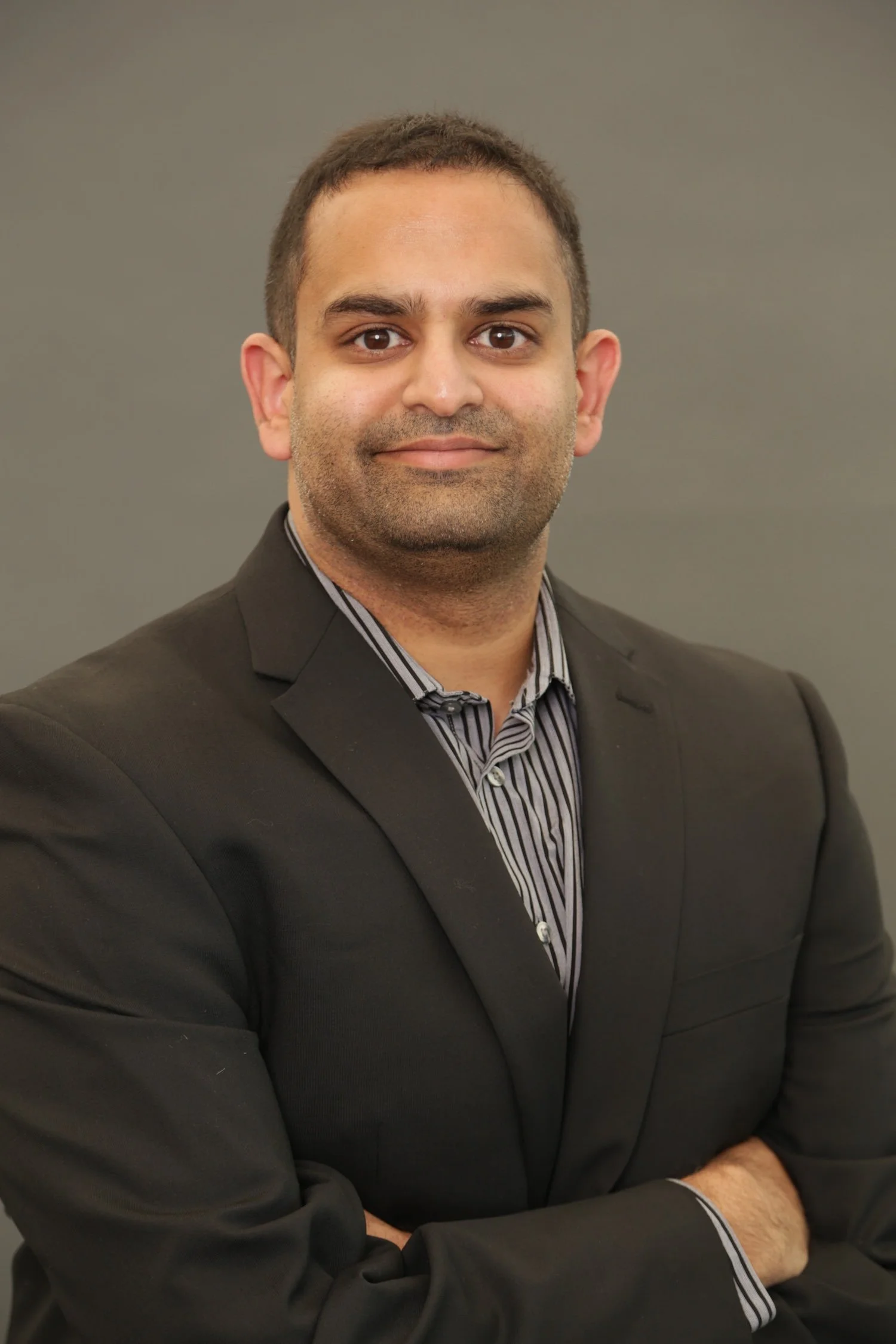A man with short dark hair, wearing a black blazer and a striped shirt, standing with arms crossed against a grey background.