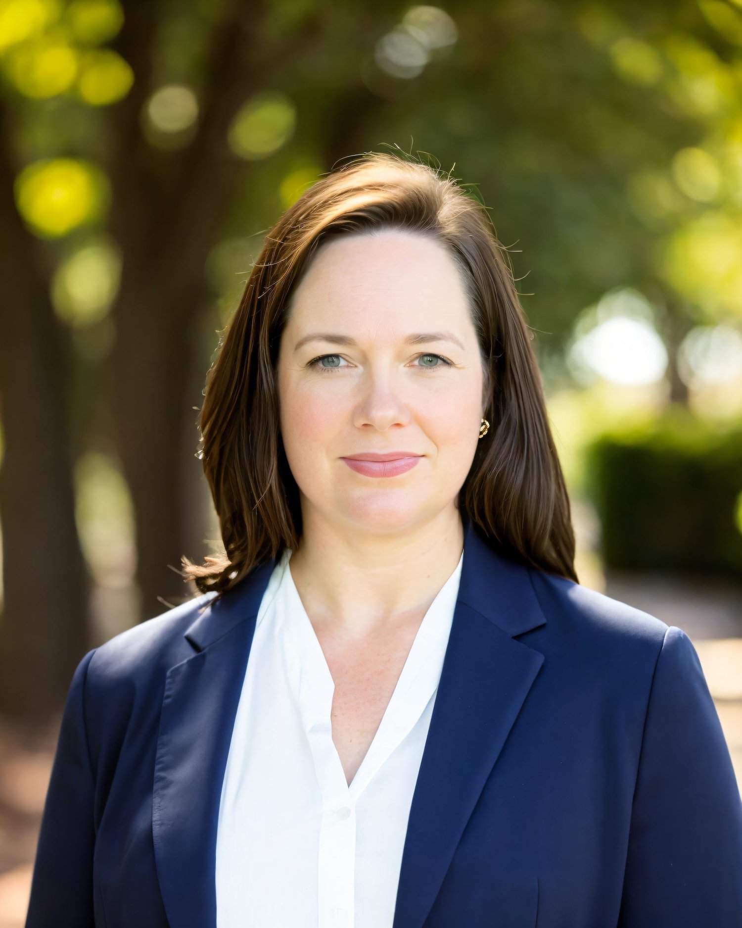 Professional woman with shoulder-length brown hair, wearing a navy blazer and white blouse, standing outdoors with blurred trees and green foliage in the background.