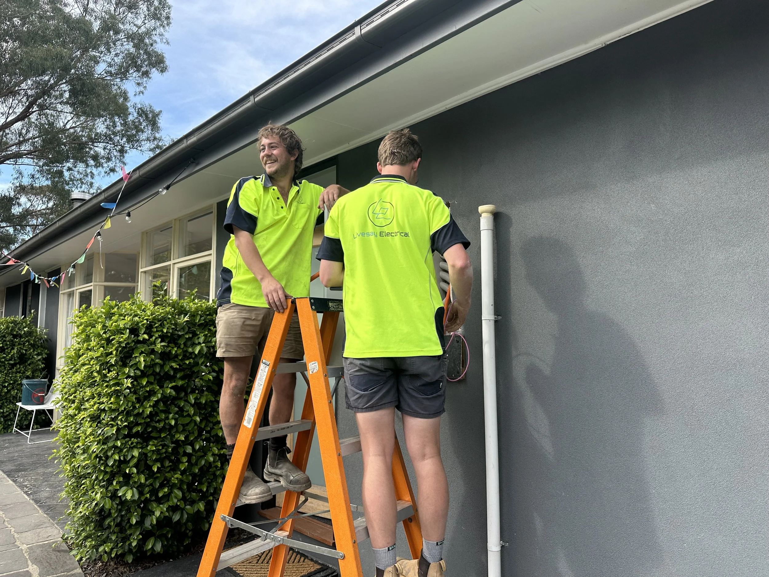 Two men in safety shirts working on electrical wiring outside a house, one on a ladder and the other standing, with a gray wall and a window in the background.