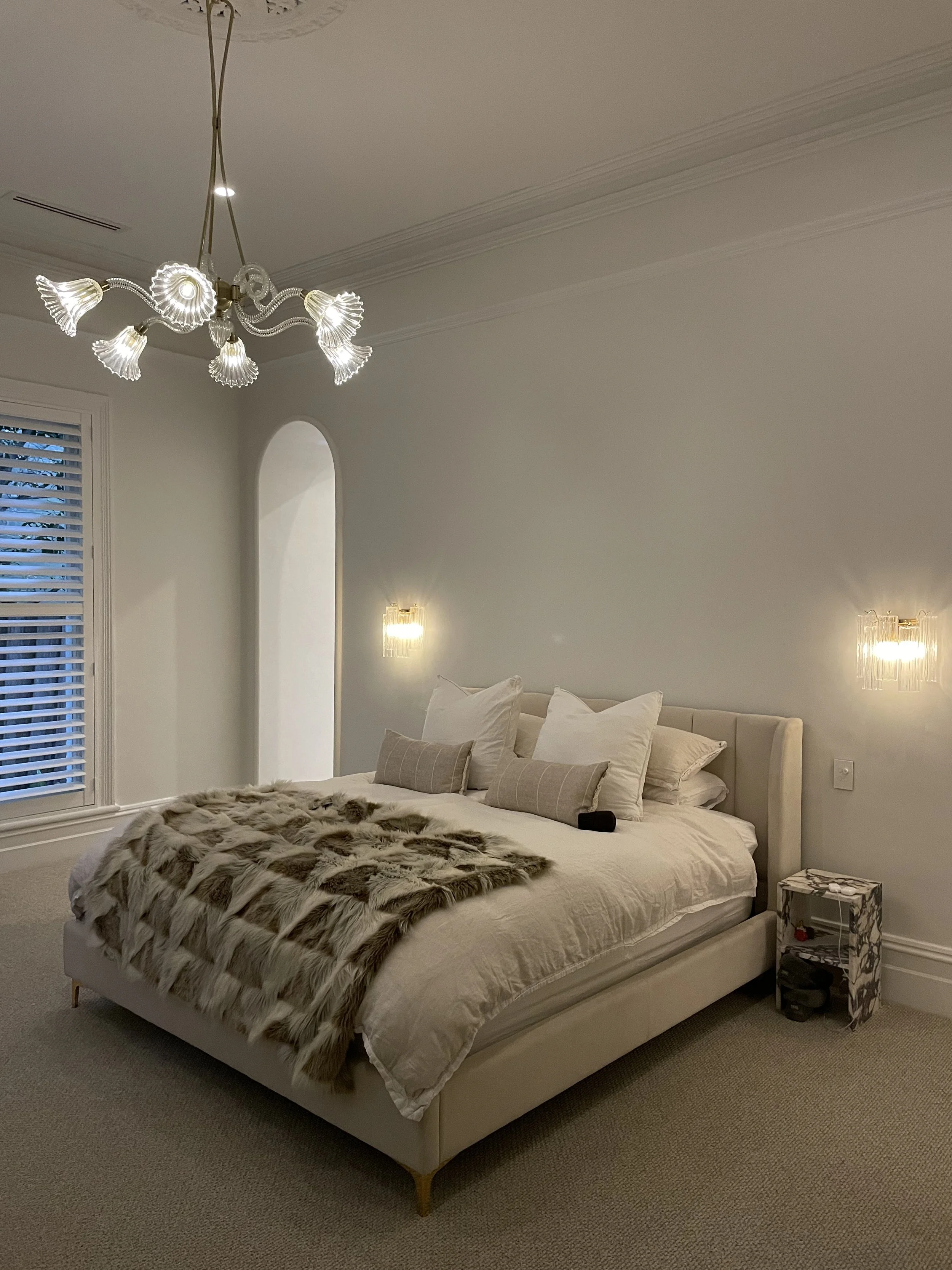 A cozy bedroom with a white bed, pillows, and a fur blanket. The room has a chandelier, wall lamps, a window with blinds, and a small nightstand.