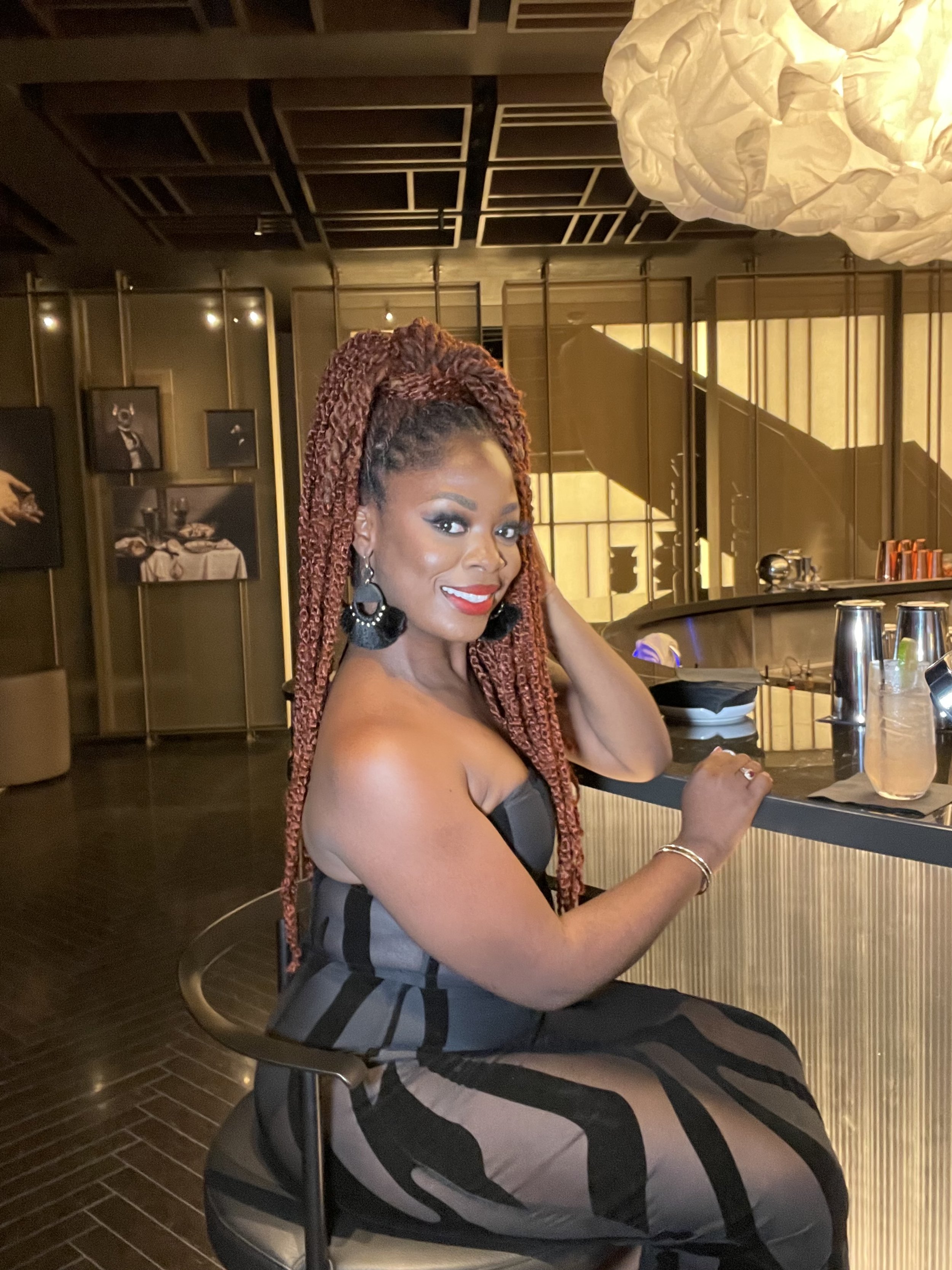A woman with styled reddish braids seated at a bar, smiling at the camera, in a modern, dimly lit restaurant or bar setting with artwork on the walls.