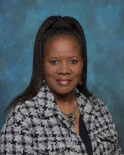 A professional woman with braided hair wearing a patterned blazer and a necklace, posed against a blue textured background.