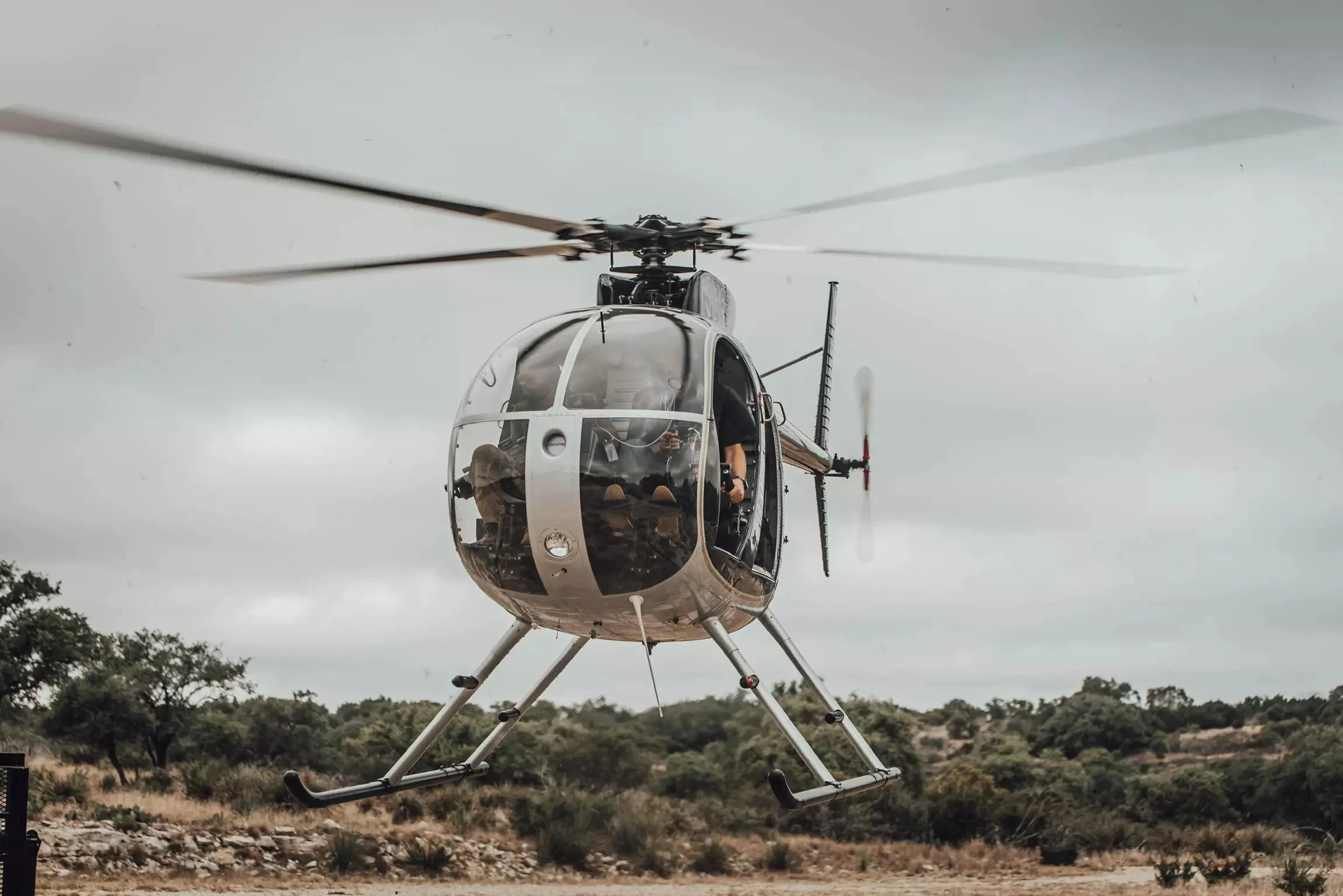 A helicopter flying close to the ground over a grassy and bushy landscape on a cloudy day.