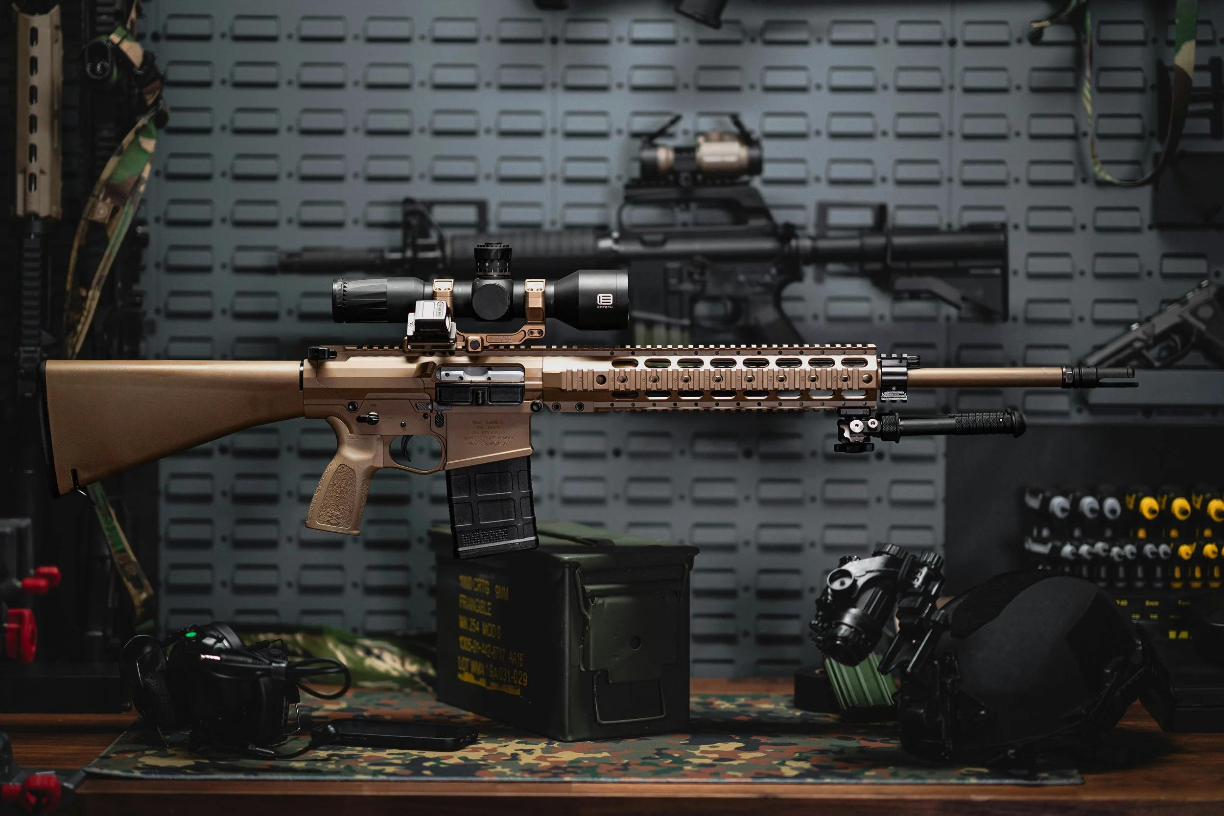 A tan-colored scoped rifle displayed on a camouflage table with firearms and tactical gear around it in a display or workshop setting.
