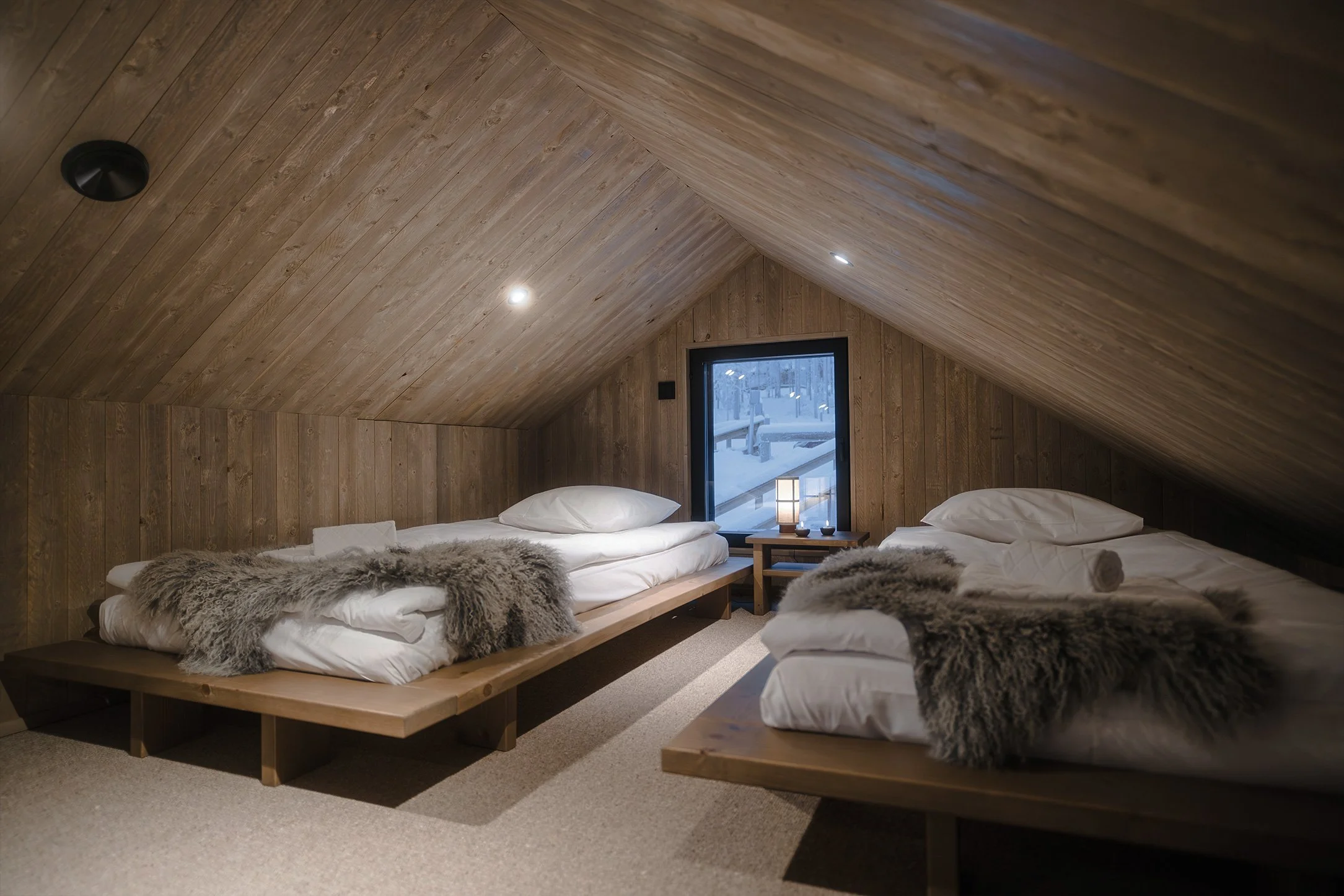 Two beds in an attic bedroom with wood-paneled walls, each bed covered with white sheets and gray fur throws. A small window with snowy outdoor view, and a nightstand with a lamp between the beds.