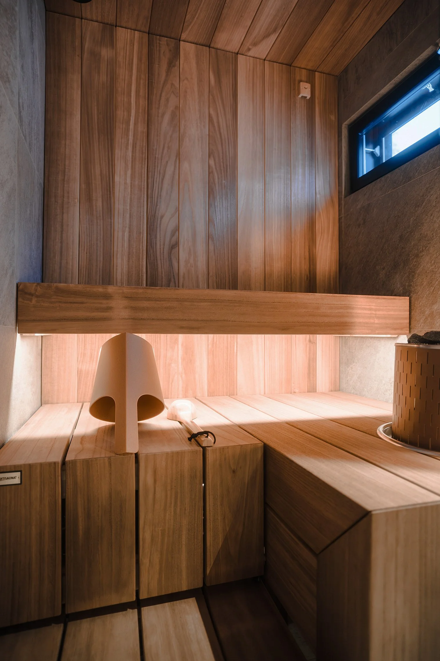 Interior of a small wooden sauna with a window, a lamp, and a rock on a basket.
